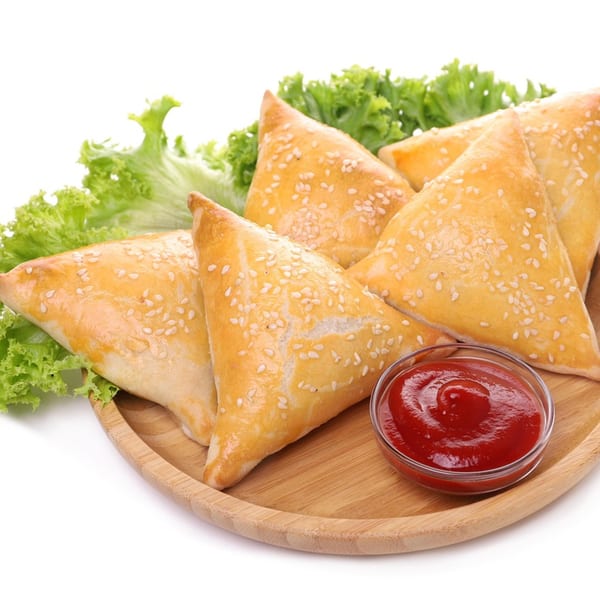 Golden-brown Indian samosas with sesame seeds, lettuce, and ketchup on a wooden plate.